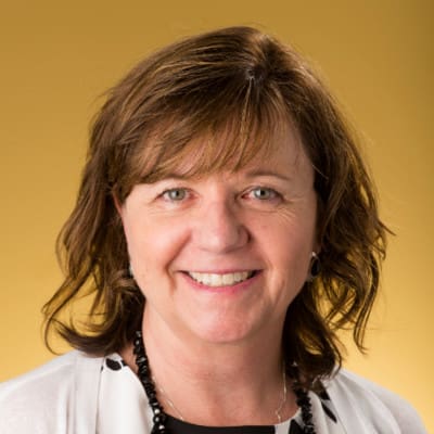 Smiling person with wavy, shoulder-length brown hair against a solid yellow background, wearing a white shirt with black polka dots, a black necklace, and a white cardigan.