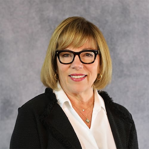 A person with shoulder-length blond hair and glasses is smiling. They are wearing a white blouse and a dark blazer, in front of a plain, textured gray background.