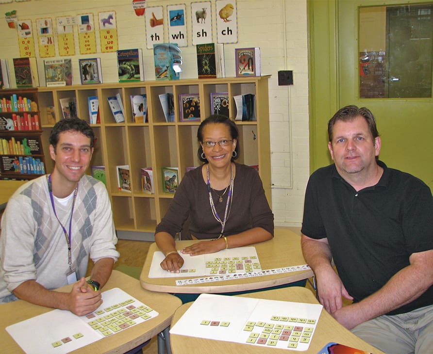 Three adults, two men and one woman, are sitting at a table in a classroom, smiling at the camera. They have educational materials on the table, such as alphabet cards. Bookshelves filled with books and educational posters are visible in the background.