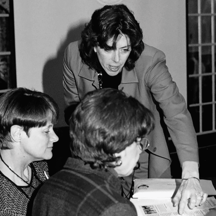 Three women are engaged in discussion at a table. One woman, standing, is pointing at a document or open book while talking to the two seated women. They are in a room with windows in the background. The image is in black and white.