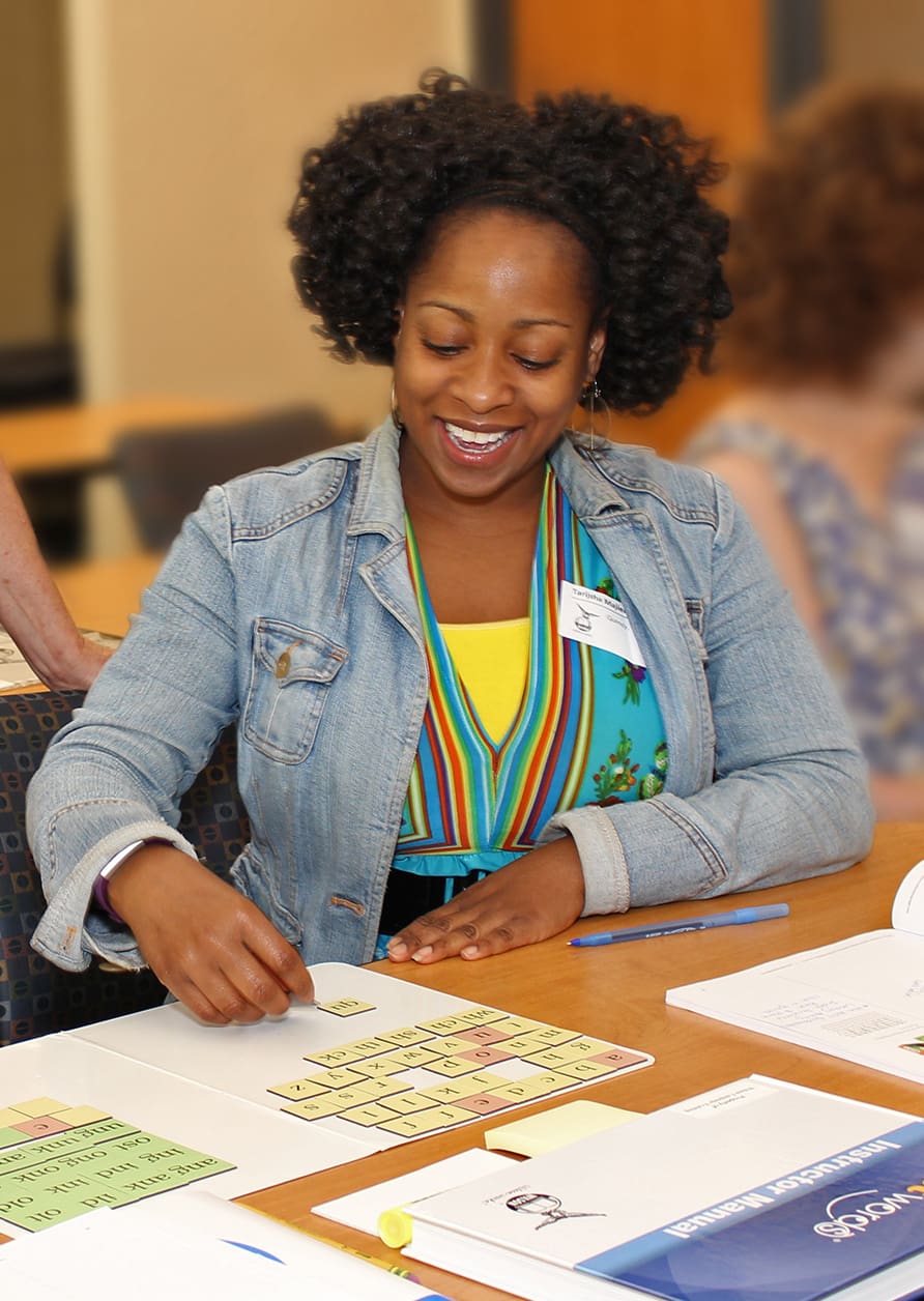 A woman with curly hair, wearing a denim jacket and a colorful striped shirt, smiles while engaging in an educational activity at a table. Various papers, a pen, and a book are spread out in front of her. Another person is partially visible in the background.