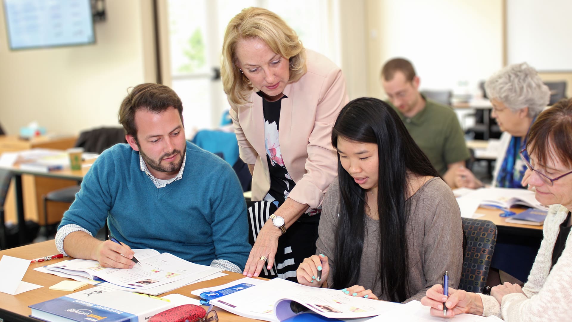 A woman stands over two seated students, assisting them with their work at a table. The students are engaged, looking at their papers, with pens in their hands. In the background, other students are also absorbed in their tasks in a well-lit classroom.