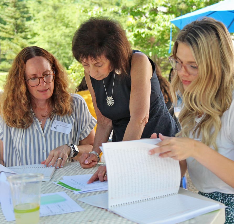 Three women are gathered around a table outdoors, engaged in a discussion while looking at documents. They appear to be collaborating on a project. One woman is standing, leaning over the table, while the other two are seated, reviewing the paperwork.