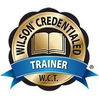 A gold circular badge with the words "Wilson Credentialed" around the top edge and "W.C.T" at the bottom. An open book is at the center, and a blue ribbon with the word "Trainer" spans the lower part of the badge.