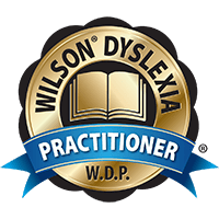 Gold badge with an open book at the center, surrounded by the words "Wilson Dyslexia Practitioner" and the initials "W.D.P." A blue ribbon with the word "Practitioner" runs across the badge.