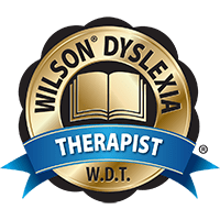 A gold badge displaying a book in the center with the words "Wilson Dyslexia" arched above and "Therapist" on a blue ribbon below. Letters "W.D.T." are at the bottom.
