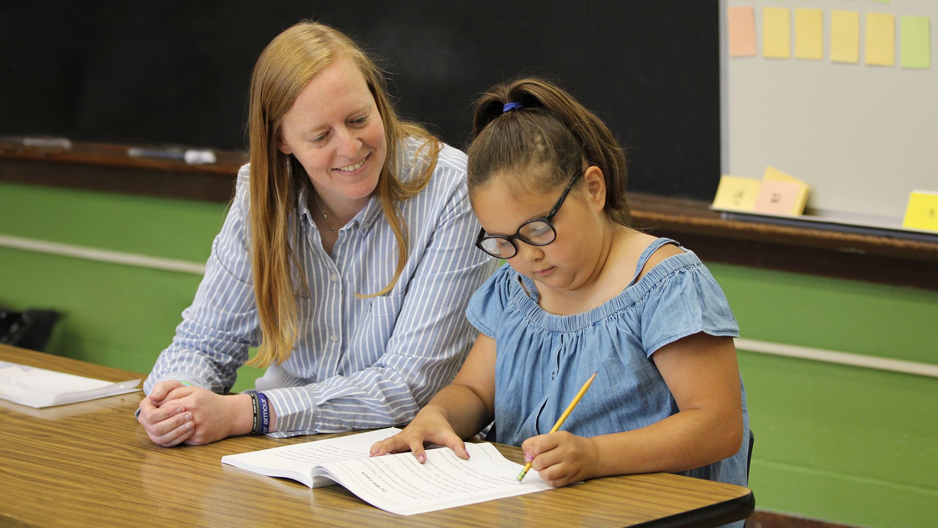 A young girl with glasses writes in a workbook at a wooden table, guided by a smiling woman sitting beside her. They are in a classroom with a blackboard and sticky notes on the wall in the background.