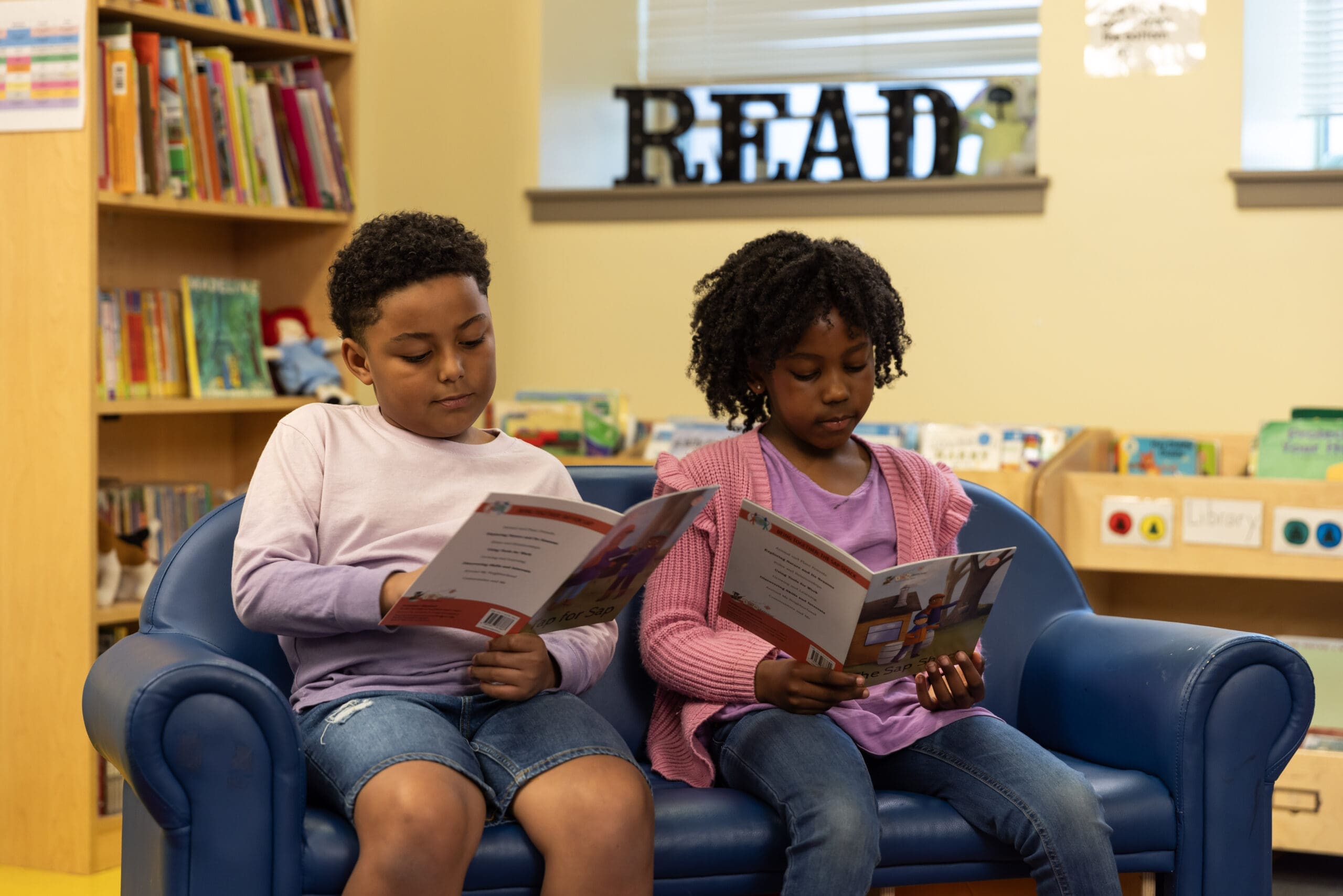 Two children sit on a blue couch in a library, reading books. Shelves filled with books and a decorative READ sign are in the background. The setting is cozy and inviting.