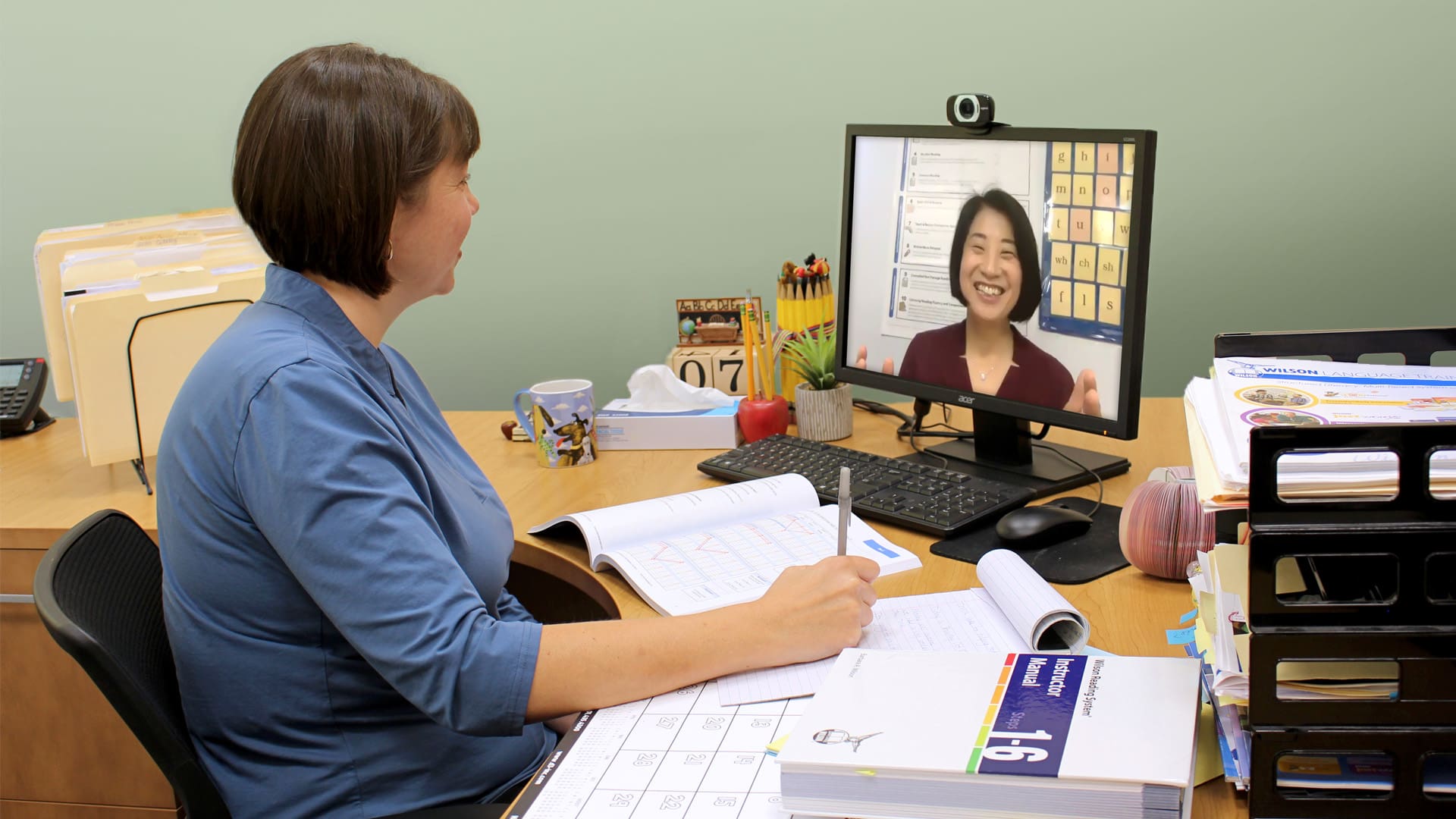 A woman sits at a desk, taking notes while having a video call on a computer. The screen shows another woman smiling. The desk has books, a calendar, a mug, and office supplies.