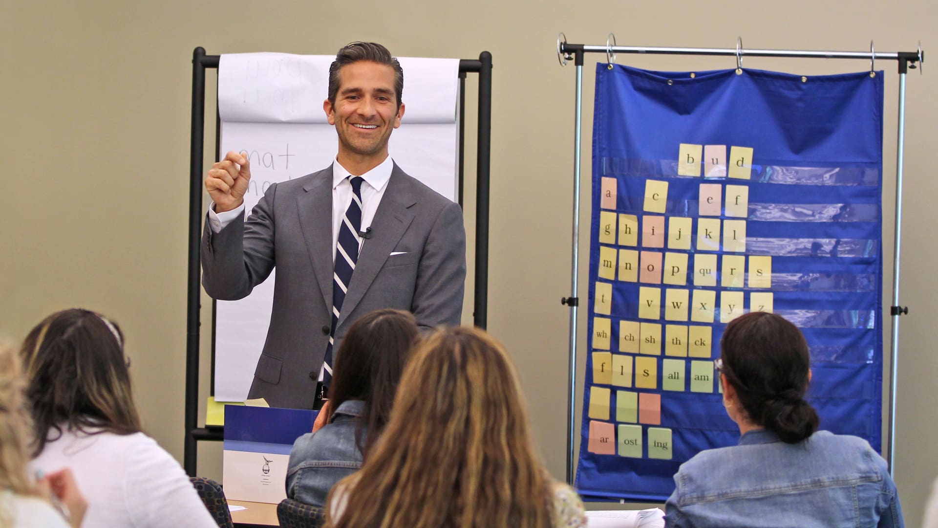 A man in a suit is standing in front of a classroom, speaking to a group of seated adults. Behind him is a flip chart and a blue board with sticky notes. The attendees are focused on his presentation.