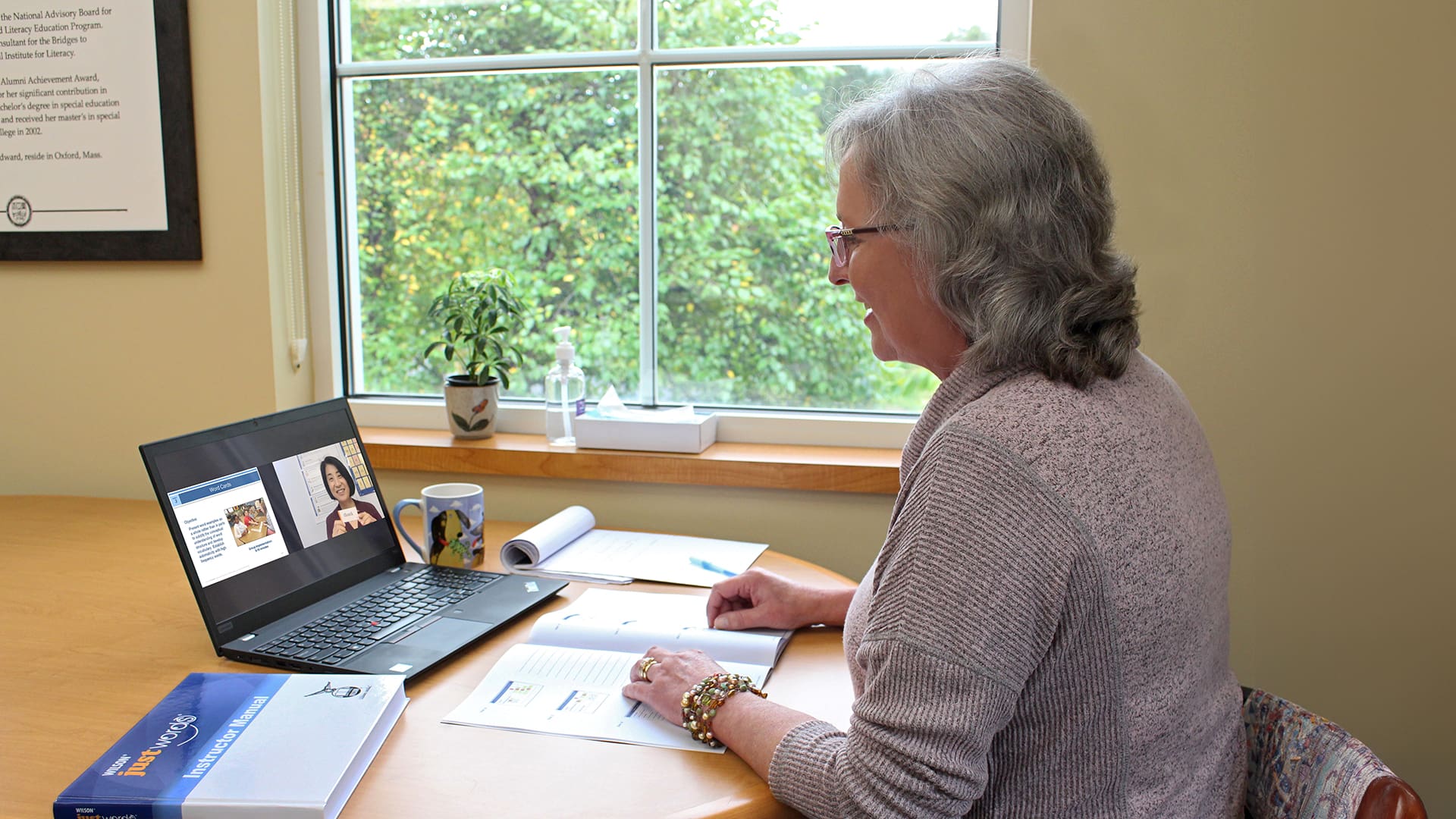 A woman with gray hair sits at a desk in front of a laptop, participating in a video call. Open books and documents are on the desk. A window with a view of trees is in the background, next to a small potted plant.