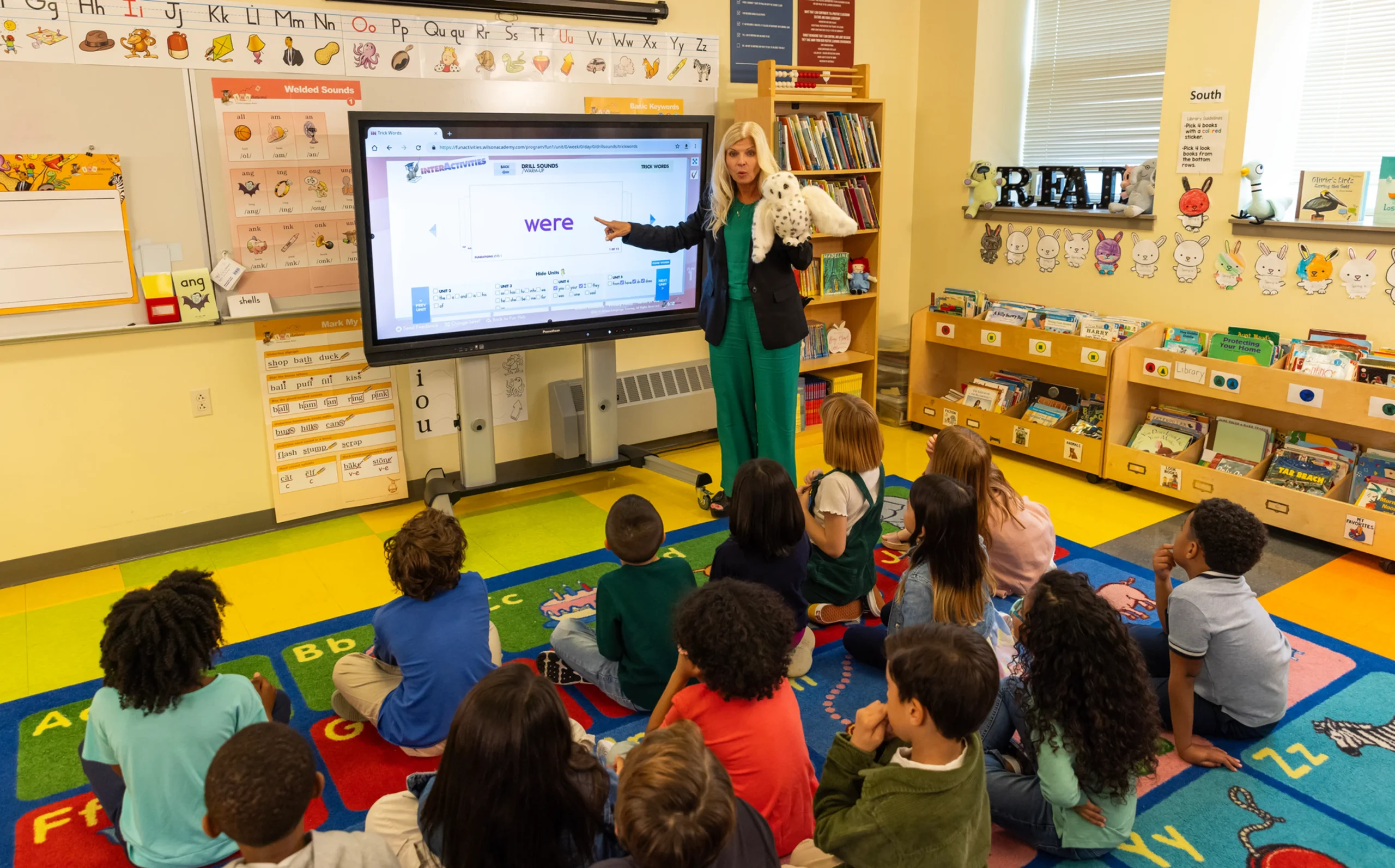 A teacher is standing in front of a screen displaying the word were, teaching a group of young children seated on a colorful carpet in a classroom. The room is decorated with books, posters, and alphabet charts.