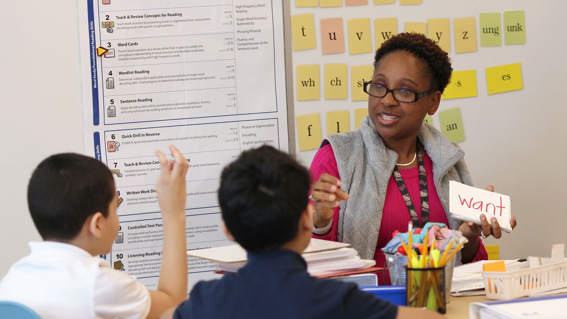A teacher sits at a table with two students, holding a flashcard. The wall behind them has educational posters and letter cards. The students are engaged, raising their hands. Various teaching materials are on the table.
