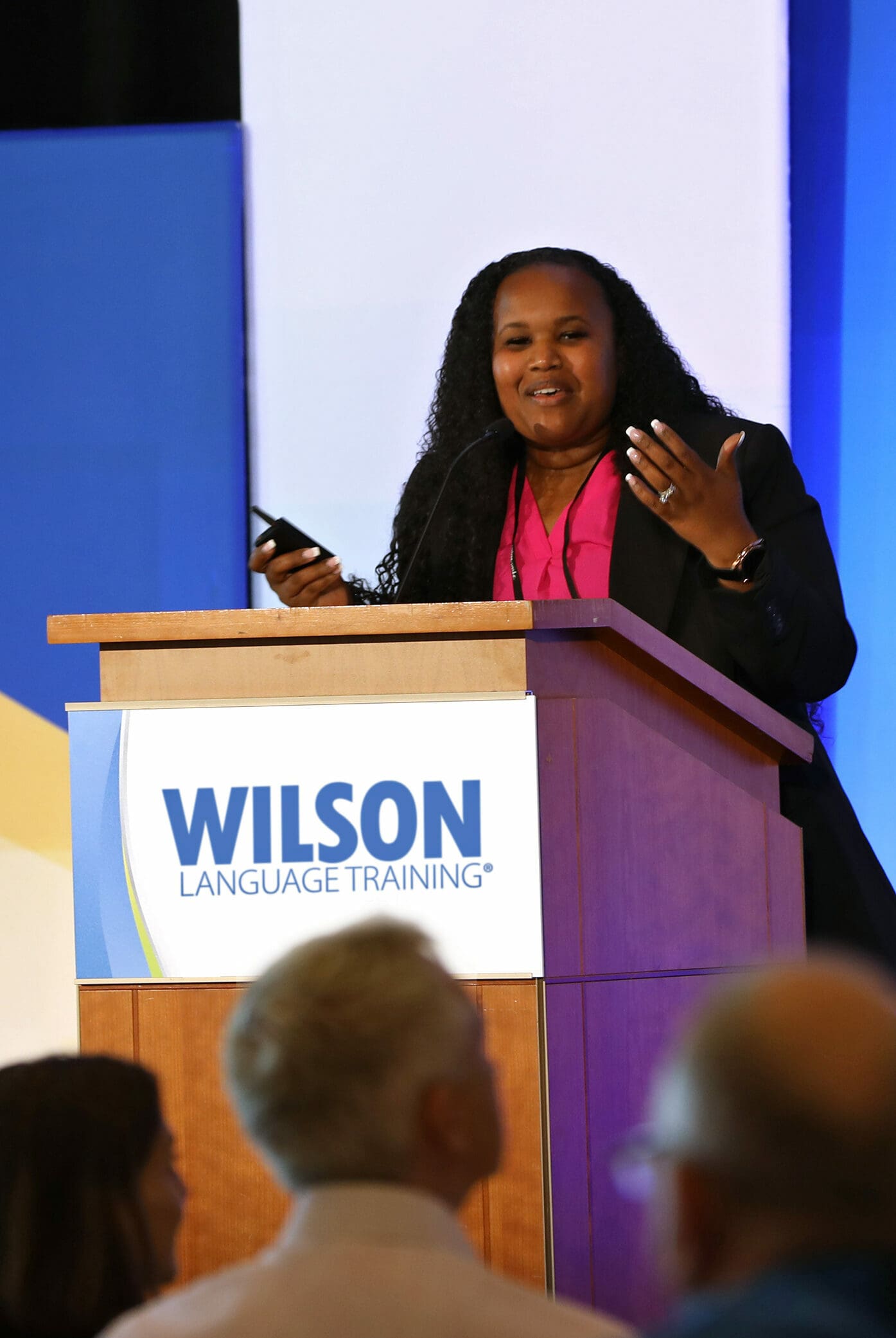 A woman with long, curly hair stands at a wooden podium, speaking and gesturing with her hand. The podium has a sign that says Wilson Language Training. She appears to be giving a presentation. People are seated in the audience, facing her.