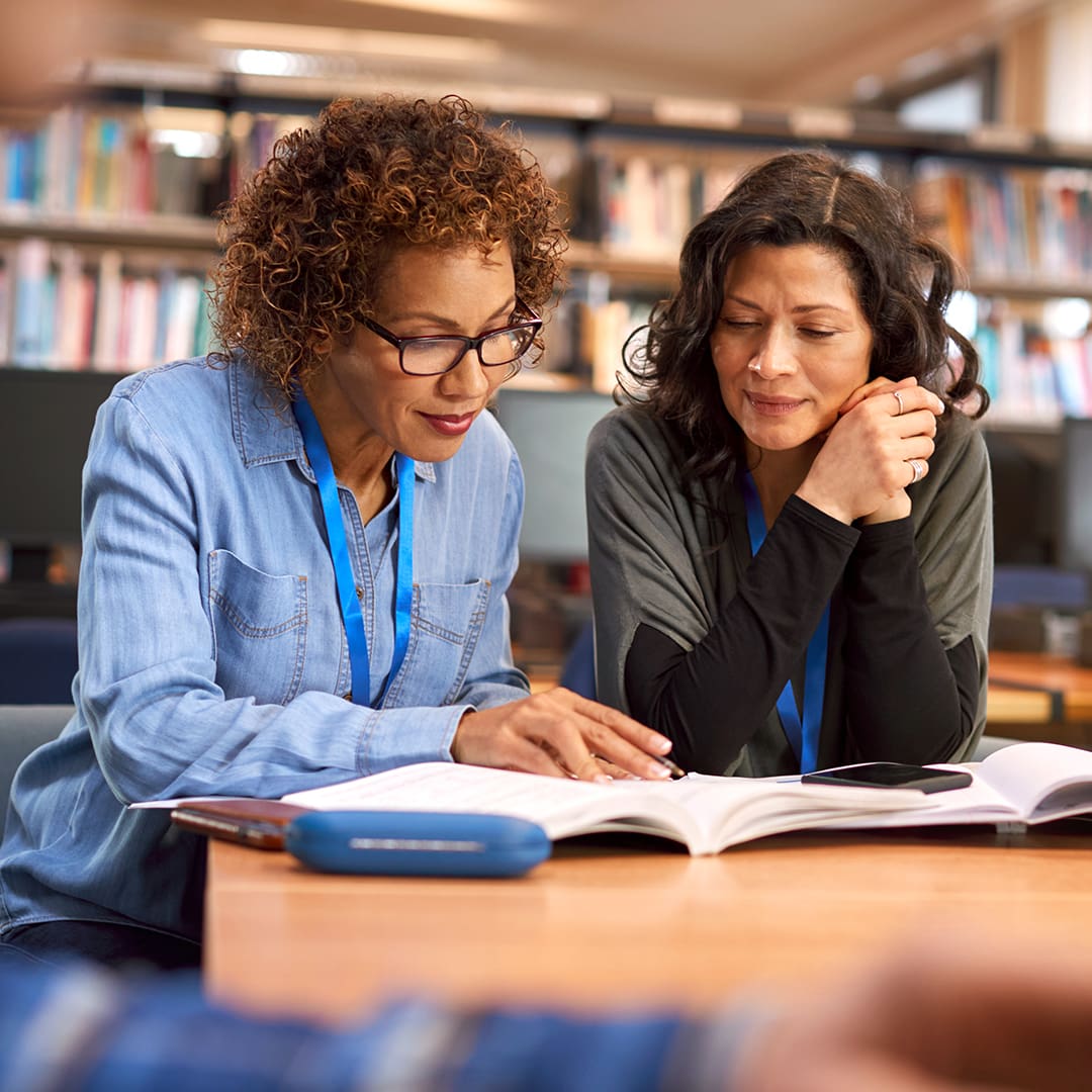 Two women are sitting at a table in a library, closely reading a book together. Both are wearing lanyards, suggesting they might be working or studying. Bookshelves filled with books are visible in the background. One woman is pointing at the book.