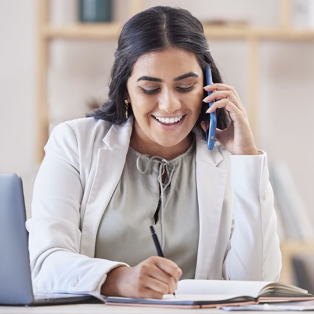 A woman wearing a white blazer and grey blouse is smiling while talking on the phone. She is seated at a desk, writing in a notebook with a pen. A laptop and books are visible on the desk. The background shows a blurred bookshelf.