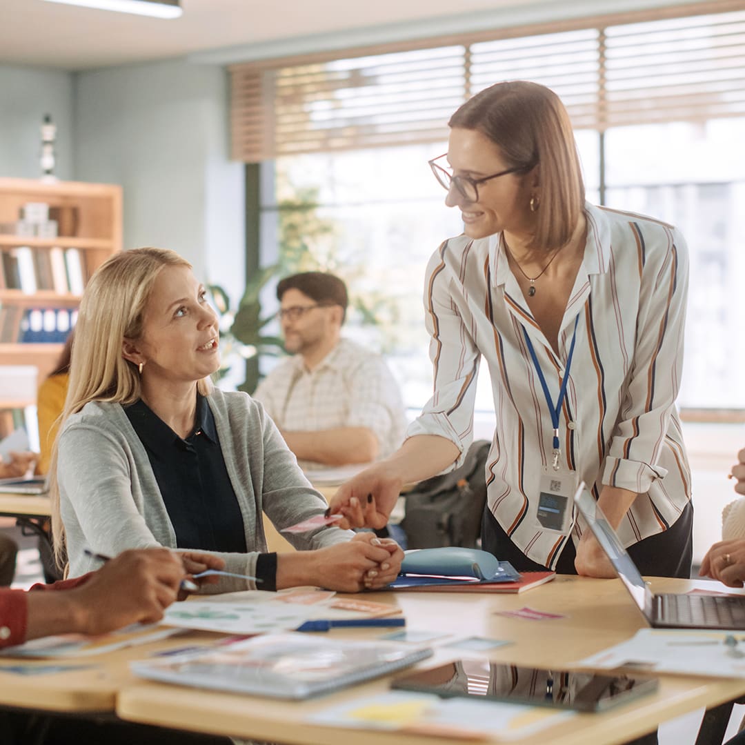 A teacher in a striped shirt and glasses engages with an attentive woman at a roundtable discussion in a bright classroom. Laptops, papers, and colorful supplies are spread out on the table. Other participants can be seen engaging and working in the background.