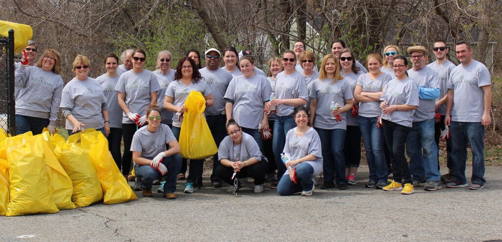 A group of people, all wearing matching gray t-shirts, pose for a photo during a community cleanup event. Some hold yellow garbage bags filled with trash, while others have gloves and trash picker tools. Trees and bushes are in the background.