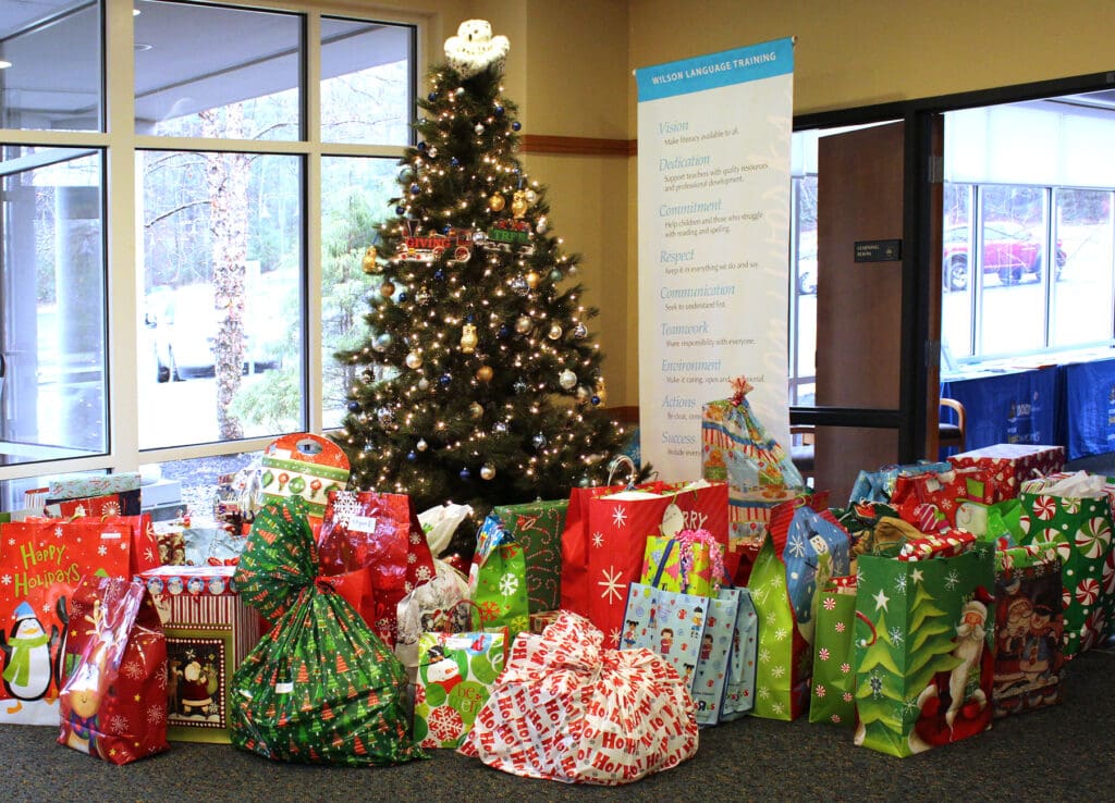 A decorated Christmas tree stands in an office lobby surrounded by a large number of wrapped holiday presents in various festive designs and colors. In the background, a banner with text and large windows are visible, showcasing a green landscape outside.