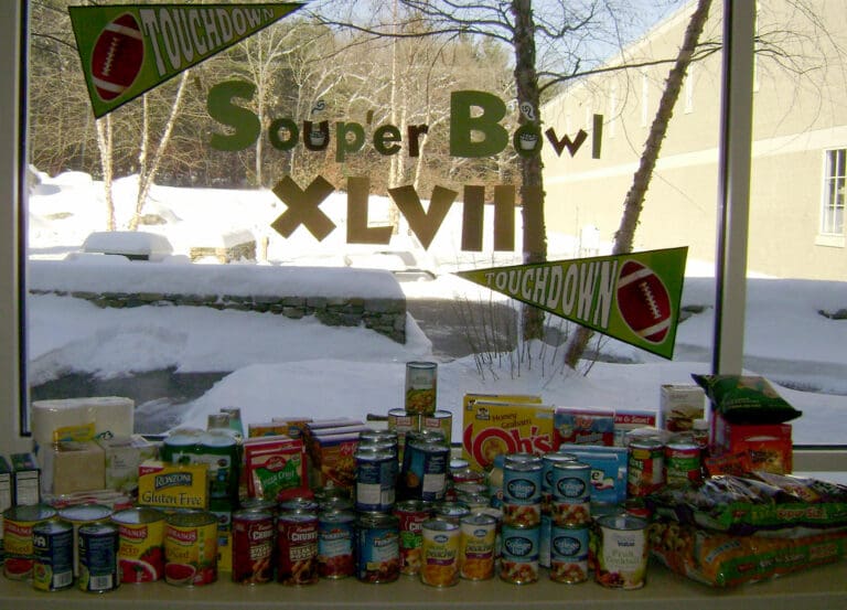A table by a window is filled with canned goods and non-perishable food items collected for a food drive. In the background, snow is visible outside. The window is decorated with banners and the text Souper Bowl XLVIII along with football-themed decals.