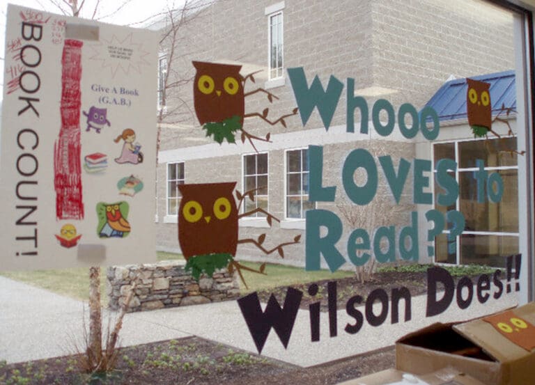 Library window decorated with colorful owls, a BOOK COUNT! poster showing 39, and the text Whooo LOVES to Read?? Wilson Does!! in large blue and green letters. Outside, a courtyard with a stone wall and windowed building is visible.