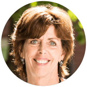A smiling woman with short, reddish-brown hair, wearing earrings and a necklace, stands in front of a blurred background with greenery.