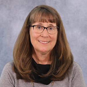 A woman with long brown hair and glasses is smiling slightly against a gray background. She is wearing a gray sweater and a black scarf.