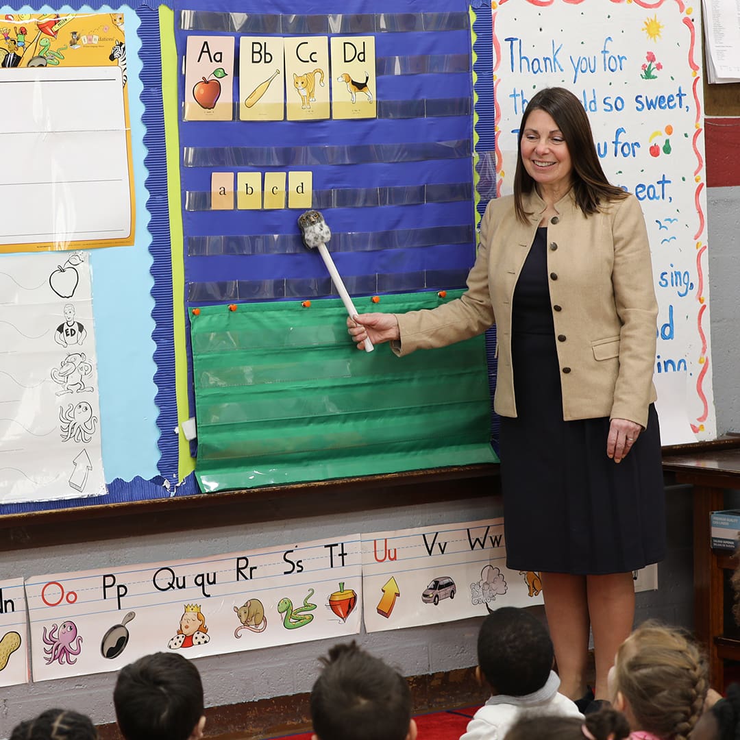 A teacher stands in front of a classroom pointing to a board with letters and pictures using a pointer. She is smiling and wearing a beige blazer over a black dress. A group of young students is seated on the floor facing her, paying attention.