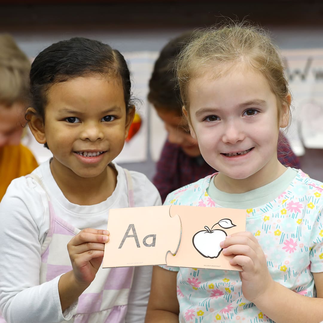 Two young girls are smiling and holding up puzzle pieces with the letter A and a drawing of an apple. They are standing in a classroom, with other children and educational materials in the background.