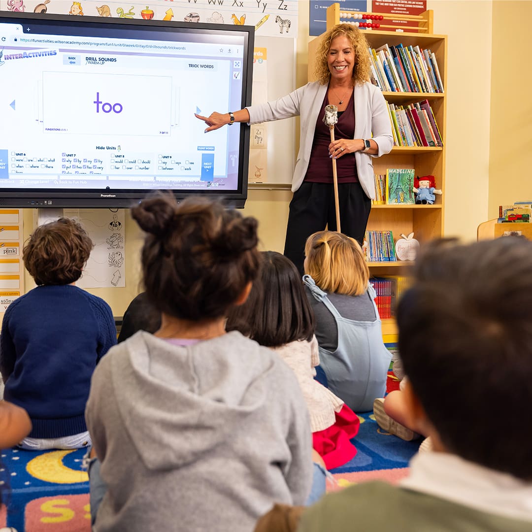 A teacher stands in front of a digital screen pointing to the word too displayed on it. She is addressing a group of young children seated on a colorful rug, with bookshelves in the background. The children are attentively watching and listening to her.