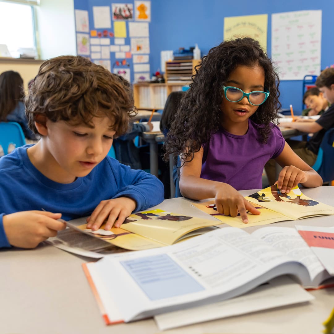 Two children are reading books at a desk in a classroom filled with other students. The boy on the left has curly brown hair and blue long sleeve shirt, while the girl on the right has long curly dark hair, glasses, and a purple shirt, pointing at her book.