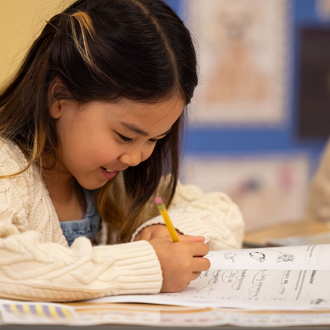 A young girl with long dark hair and a beige sweater smiles while writing with a pencil in a workbook. She is seated at a desk in a classroom with colorful bulletin boards in the background.
