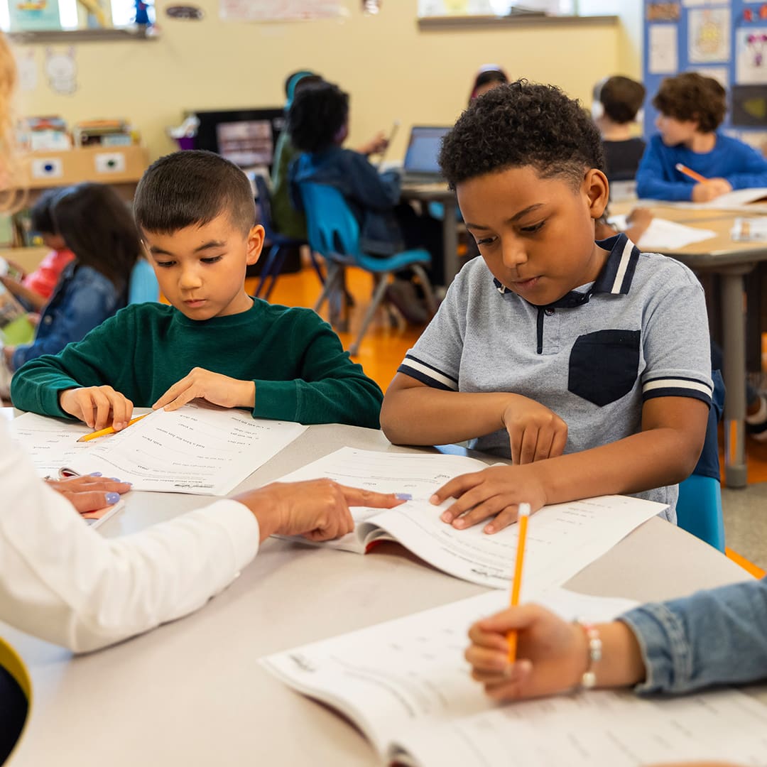A diverse group of young students sits at a table in a classroom, working on worksheets. Two boys are prominently focused on their papers while a teacher points to one of the boy's work, providing guidance. Other children and desks with laptops are visible in the background.