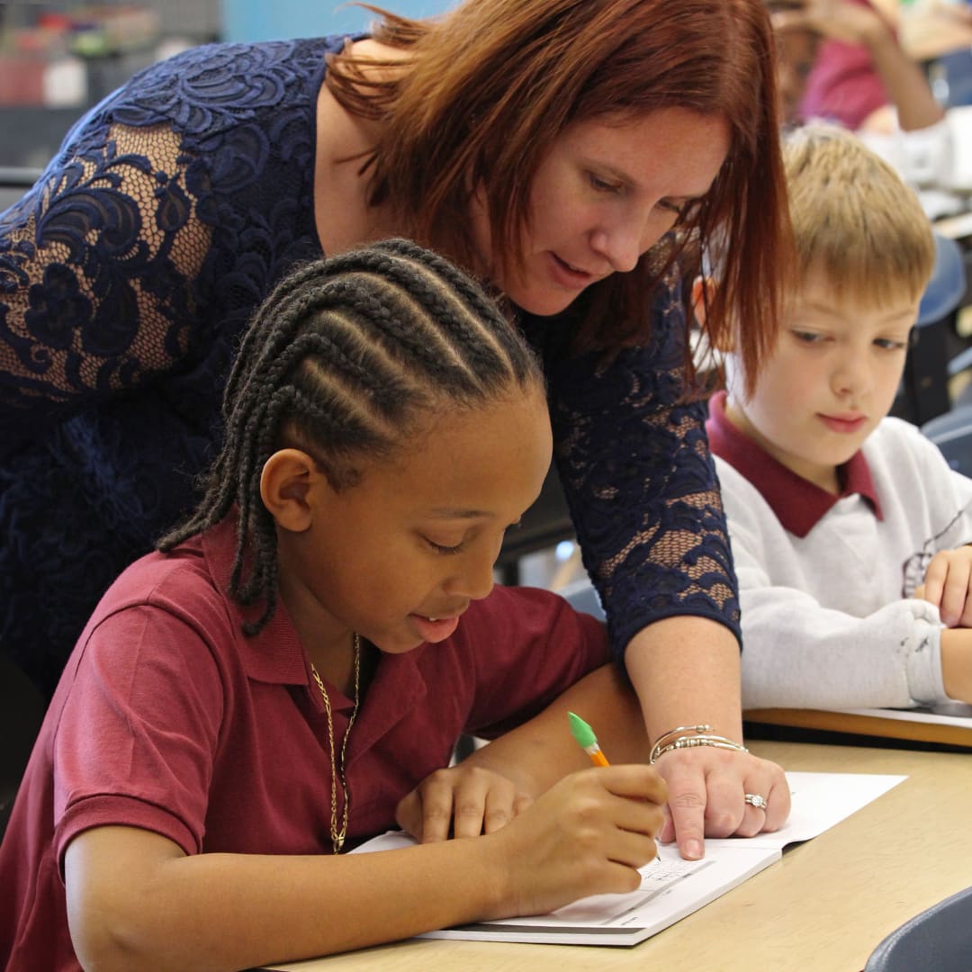 A teacher with red hair leans over to help a young student with braids who is writing in a notebook. Another student with light hair looks on. They are seated at a desk in a classroom.