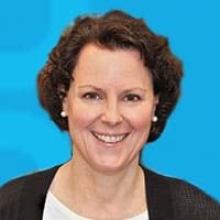 A person with short, curly hair and pearl earrings smiles against a bright blue background. They are wearing a white top and a dark cardigan.