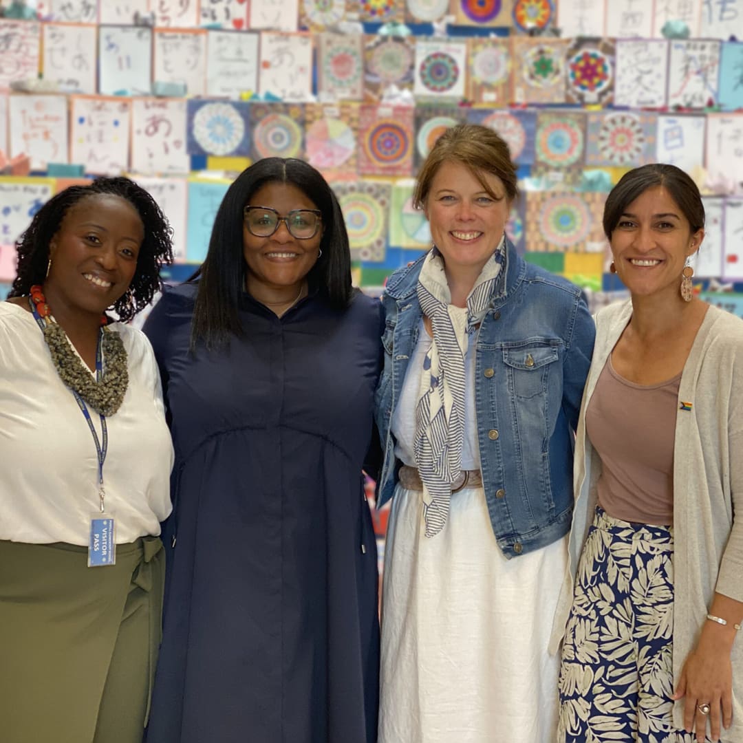 Four women are standing side by side, smiling at the camera. They are in front of a colorful background consisting of various drawings and artwork. The women are dressed in casual to semi-formal attire.