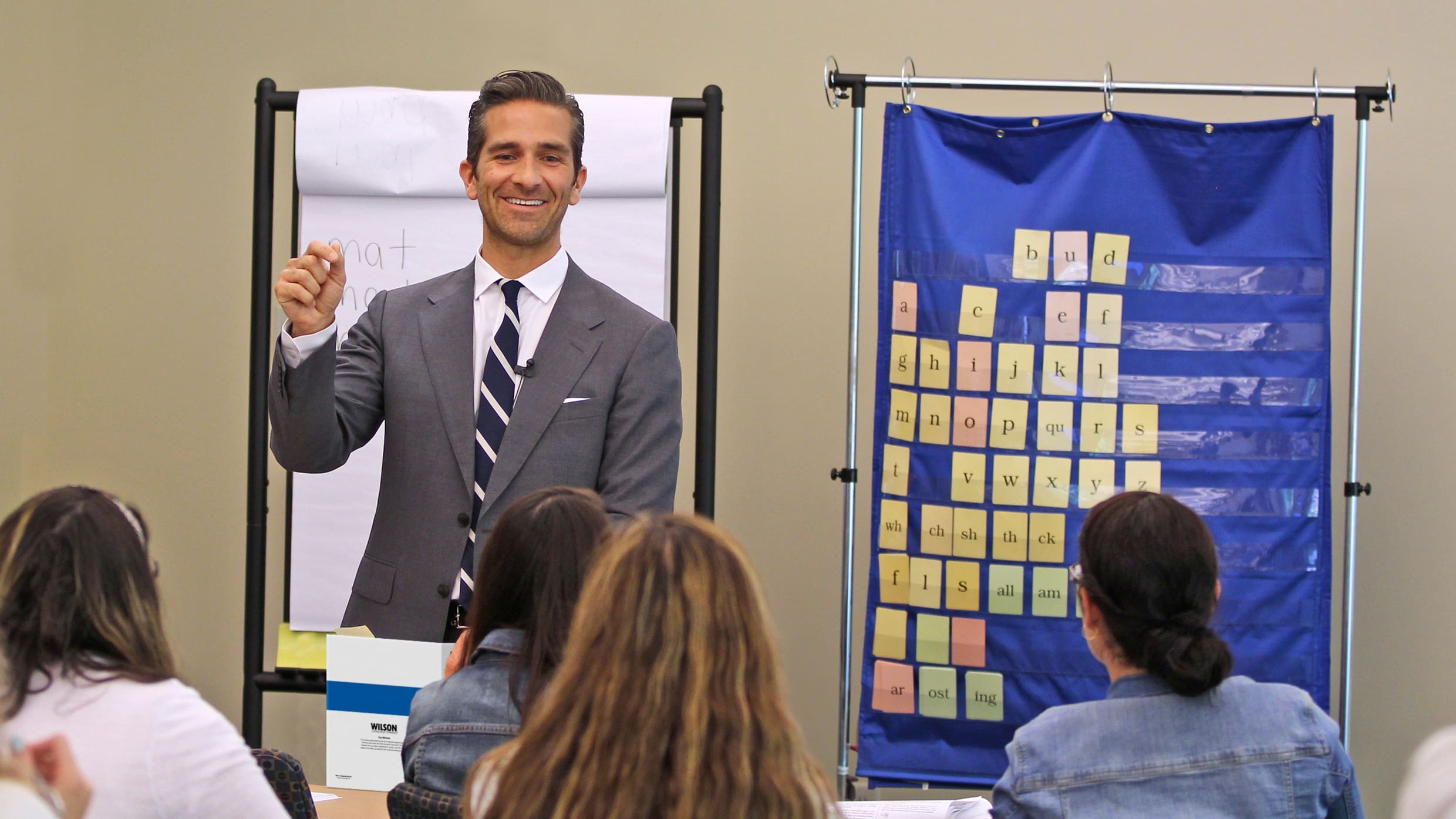 A man in a suit stands in front of a group of people, pointing at a flip chart and smiling. A blue chart with letters on sticky notes is hanging on the wall beside him. The attendees are seated, facing him, and appear to be paying attention.