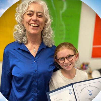 A smiling woman with curly gray hair and a blue shirt stands next to a girl with glasses and long hair, holding a Certificate of Achievement. They are indoors, with colorful art in the background.