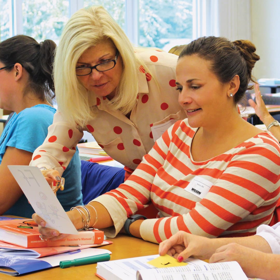 A woman with glasses and a polka dot shirt assists another woman with a worksheet. They are seated at a table with books and papers. Other people are visible in the background, engaged in similar activities.