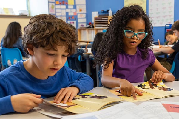 Two children sitting at a classroom table, engaged in reading books. The boy with curly hair is wearing a blue shirt, and the girl with long curly hair and glasses is wearing a purple shirt. Other students are visible in the background.