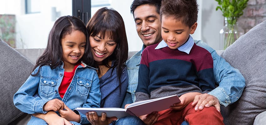 A family of four sits on a couch reading together. The mother and daughter share a book, while the father and son look at another book. They are smiling and appear to be enjoying their time together. The background is softly blurred.