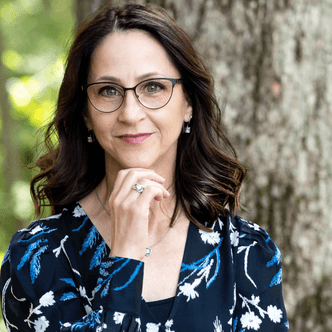 A woman with long wavy dark brown hair and glasses stands outdoors in front of a tree. She wears a navy dress with a white and blue floral pattern and rests her hand gently near her chin, smiling softly.