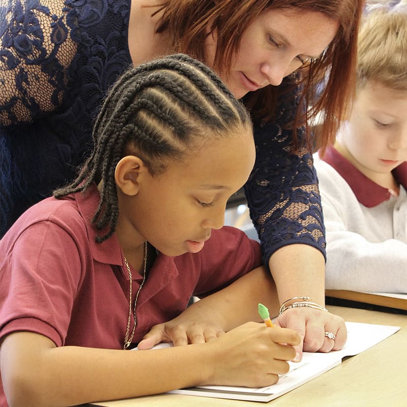 A teacher leaning over a student and pointing to the student's paper on the desk