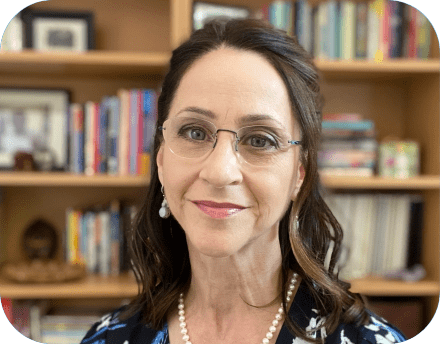 Bonnie Singer smiling in front of a bookshelf filled with colorful books and framed photos. She wears glasses, pearl earrings, a pearl necklace, and a floral-patterned top.