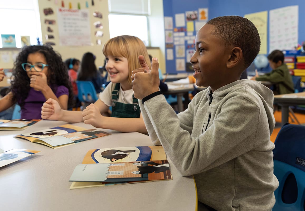 Two students with books on their desk as the give a thumbs-up