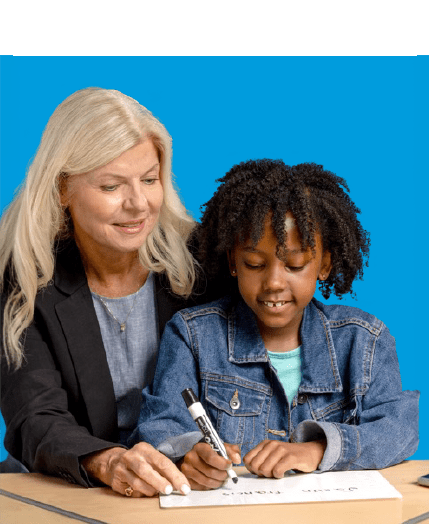 Adult woman and young girl seated at a table. The woman gently guides the girl's hand as she writes with a marker on a whiteboard.