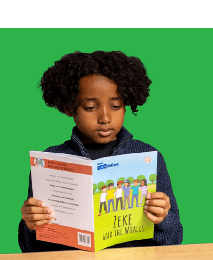 Young boy with curly hair reads a book titled "Zeke and the Whales" while seated at a desk.