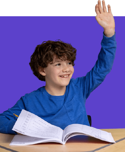Smiling boy in a blue shirt raises his hand while looking to the side, seated at a desk with an open workbook.