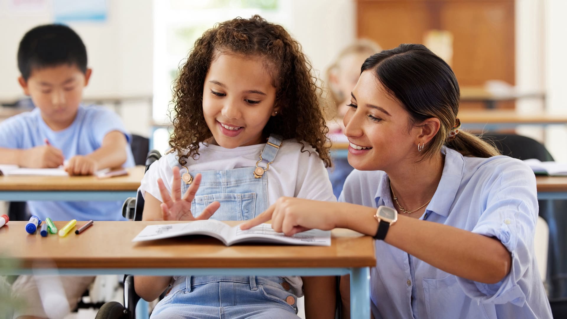 Teacher helps young student in a wheelchair with Fundations K-3 reading lesson, smiling.